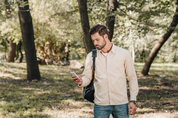κομψό άνδρα σε casual ρούχα με σακίδιο χρησιμοποιώντας το smartphone στο πάρκο - Φωτογραφία, εικόνα