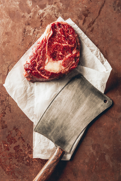 top view of raw meat steak, baking paper and cleaver on tabletop in kitchen - Foto, Bild