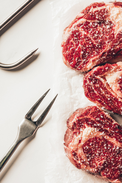 top view of three pieces of raw meat steaks with salt, meat fork and metal hook on white surface - Photo, Image