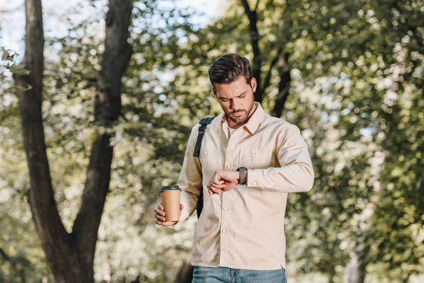 portrait of man with backpack and coffee to go checking time in park - Photo, image