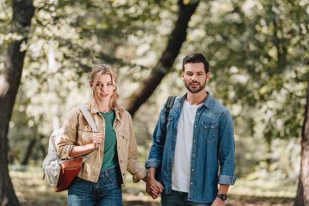 πορτρέτο του νεαρό ζευγάρι με σακίδια, κρατώντας τα χέρια και κοιτάζοντας κάμερα στο πάρκο - Φωτογραφία, εικόνα