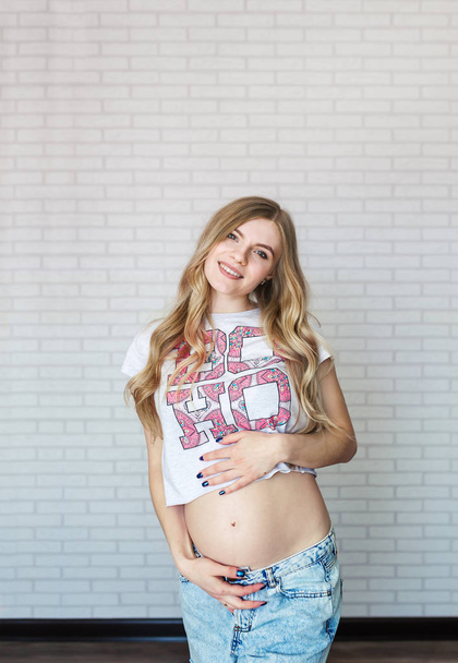 Hermosa joven embarazada con el pelo largo vestido con jeans, una camiseta blanca en el fondo de la pared de ladrillo. Retrato vertical
 - Foto, imagen