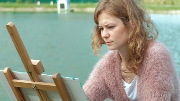 Kaunis nainen, jolla on punaiset hiukset, maalaa kuvan kankaalle, joka seisoo maalaustelineessä. Nainen on ulkona lähellä järven jokea, hän vetää elämästä
 - Materiaali, video