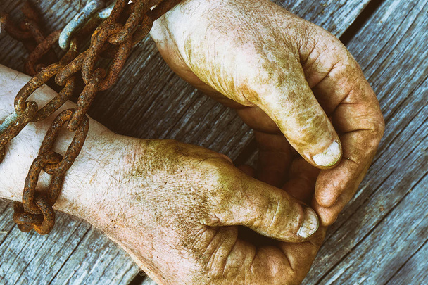Dirty male hands chained with old rusty thick chain on the wooden boards with a copy space. Unfreedom concept. - Foto, Imagen