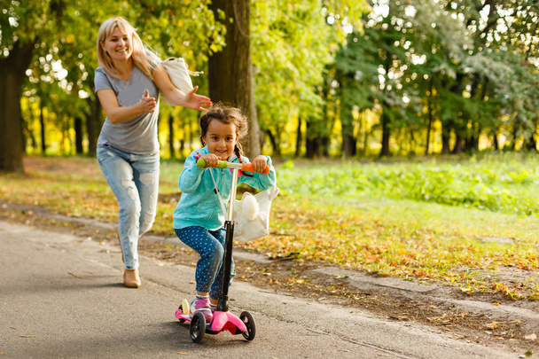 jovem mãe ensinando sua filhinha a andar de scooter no parque
 - Foto, Imagem
