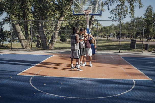 Group of young  basketball players standing on outdoor court and looking at smartphone. - Photo, Image