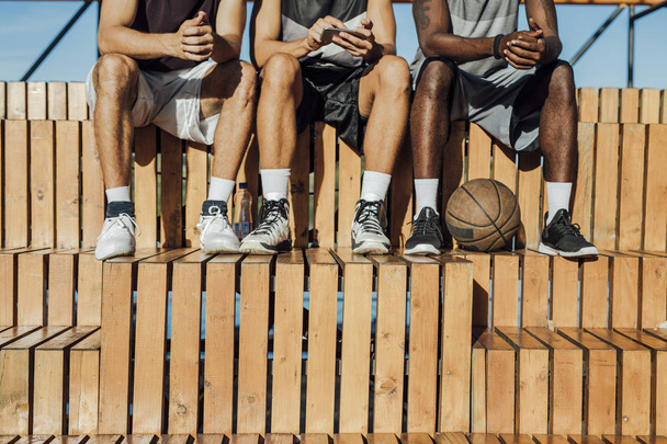 multiethnische Gruppe von nicht wiedererkennbaren Basketballspielern, die am Spielfeldrand sitzen und Mobiltelefone benutzen. - Foto, Bild