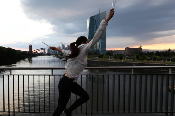 Eine junge Frau läuft mit Feuerwerkskörpern in der Hand vor einer städtischen Skyline und einem Fluss. Profilansicht. Dreiviertellänge.  - Foto, Bild