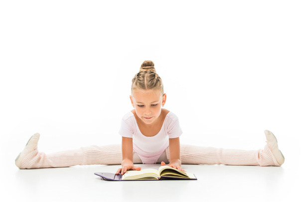 concentrated little ballerina reading book and doing twine isolated on white background  - Photo, Image