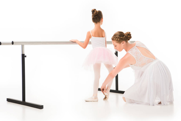 rear view of female teacher in tutu checking pointe shoes of little ballerina exercising at ballet barre stand isolated on white background  - Фото, зображення