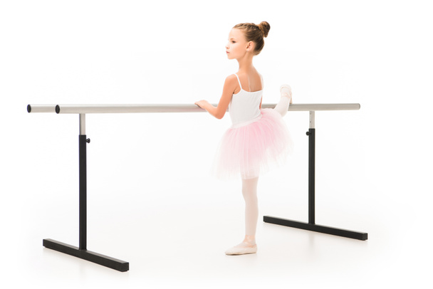 side view of little ballerina in tutu and pointe shoes practicing at ballet barre stand isolated on white background  - Zdjęcie, obraz