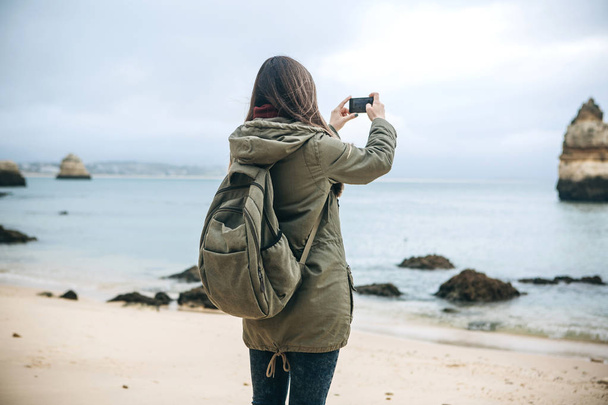 Chica turista con una mochila fotografiando un hermoso paisaje de pie en la costa atlántica
 - Foto, Imagen