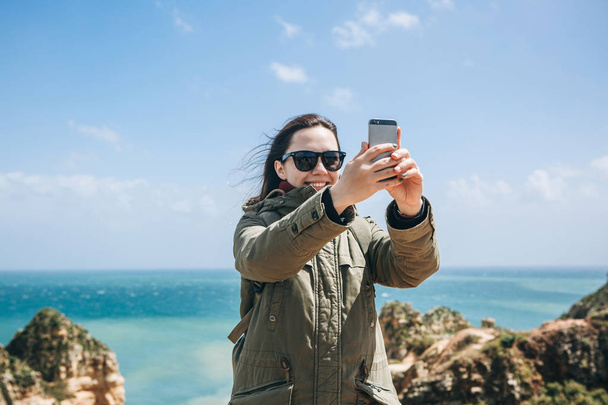 ein positives Touristenmädchen macht einen schönen Anblick oder macht ein Selfie oder kommuniziert mit jemandem online mit einem Handy - Foto, Bild