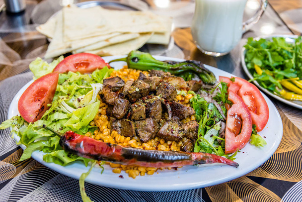 Турецкий шашлык подается на белой тарелке с нарезанным лаваш, петрушкой, луком, салатом, помидорами, жареным перцем, смешанными огурцами и айраном
. - Фото, изображение