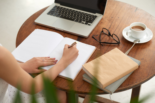 Руки студента, пишущего в дневнике ручкой за столом с ноутбуком, кофейной чашкой, книгами и стаканами
  - Фото, изображение
