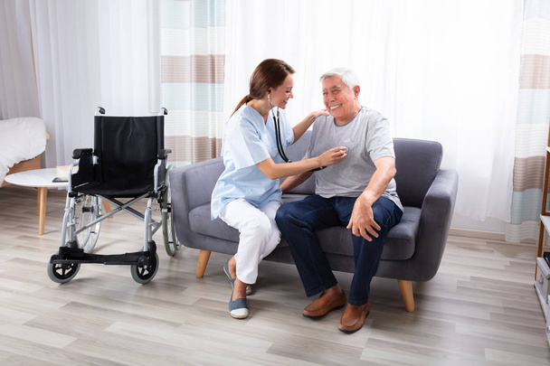 Female Nurse Checking Happy Senior Man Sitting On Sofa With Stethoscope - Фото, изображение