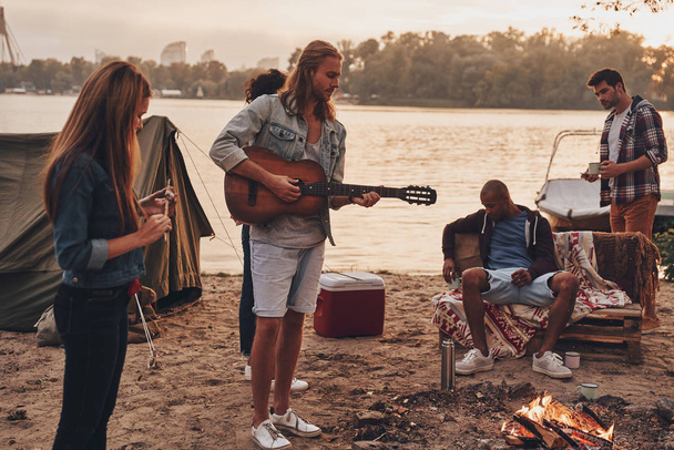 young people in casual wear enjoying camping near lake at sunset, man playing guitar  - Fotó, kép