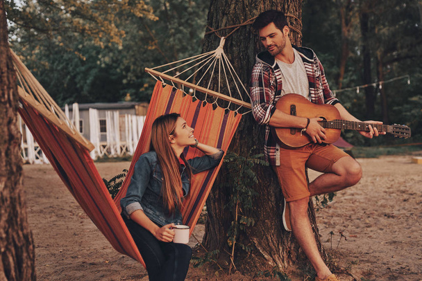 handsome young man playing guitar for beautiful girlfriend outdoors in forest, woman sitting in hammock with cup  - Fotó, kép