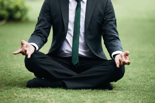 Empresário irreconhecível de terno meditando na grama verde no parque
 - Foto, Imagem