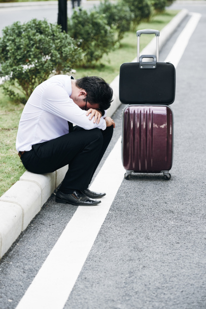 Frustrated Asian businessman sitting on border stone with his luggage bags nearby - Foto, imagen