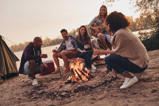 friends in casual wear smiling while enjoying beach party near campfire with guitar  - Foto, imagen