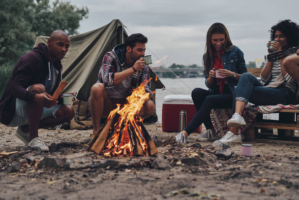 friends sitting and talking at campfire with hot drinks in cups  - Foto, imagen