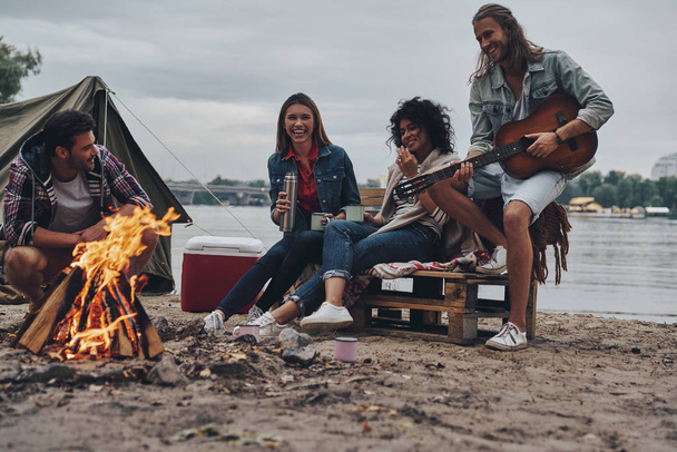 young people in casual wear enjoying camping near lake at sunset, man playing guitar  - Photo, image