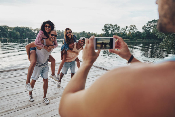 beautiful young couples spending carefree time while standing on pier, man taking mobile photo of friends having fun with piggyback - Photo, image