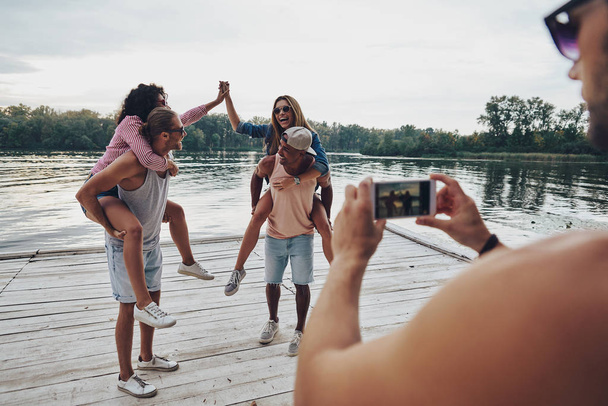 beautiful young couples spending carefree time while standing on pier, man taking mobile photo of friends having fun with piggyback - Zdjęcie, obraz