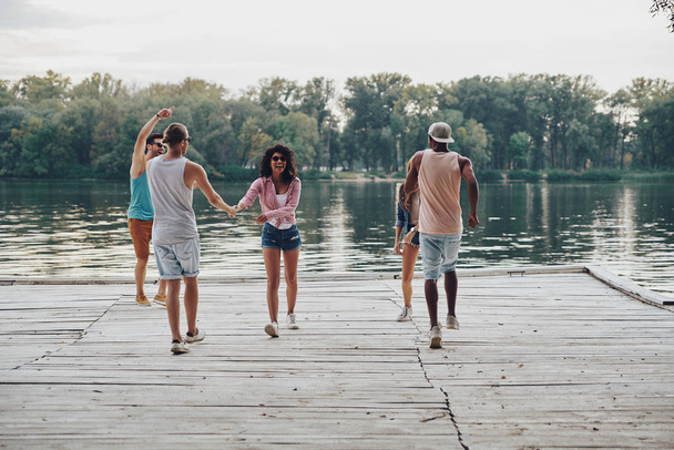 full length of young people in casual wear smiling and dancing on wooden pier at river  - Foto, Imagem