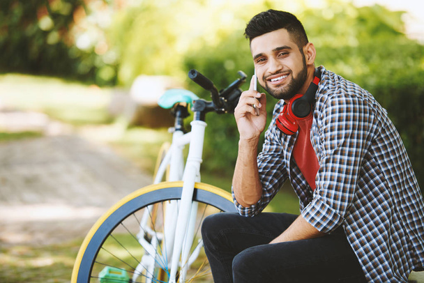 Vrolijke man op zoek naar camera met toothy glimlach terwijl praten op smartphone en zittend op een bankje, moderne fiets op achtergrond - Foto, afbeelding