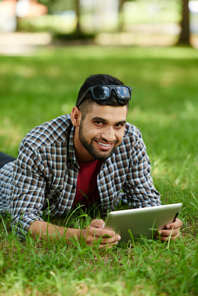 lächelnder indischer Mann, der auf grünem Rasen liegt und beim Surfen im Internet auf einem modernen digitalen Tablet für Fotos posiert, Porträtaufnahme - Foto, Bild