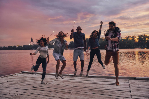 full length of young people jumping with Bengal lights on wooden pier at lake with forest and sunset sky  - Photo, Image