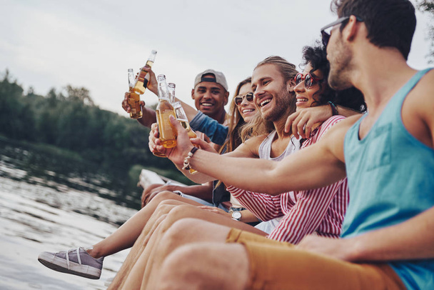 happy young people in casual wear smiling and toasting with beer while sitting on pier at river - 写真・画像