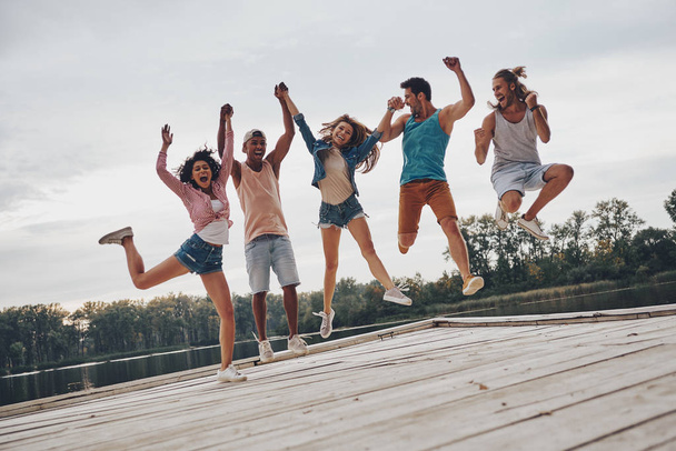 full length of young people in casual wear jumping on wooden pier at lake with forest  - Foto, Imagen