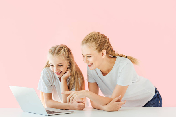 smiling mother and daughter in white t-shirts pointing and looking at laptop together isolated on pink - Photo, Image