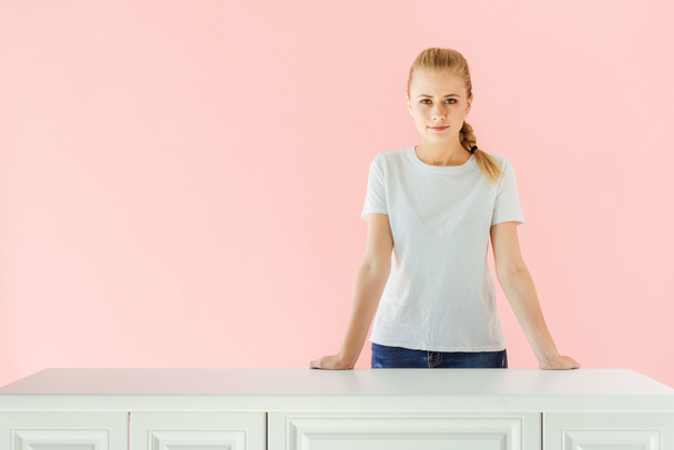 attractive young woman standing behind kitchen table isolated on pink - Photo, Image