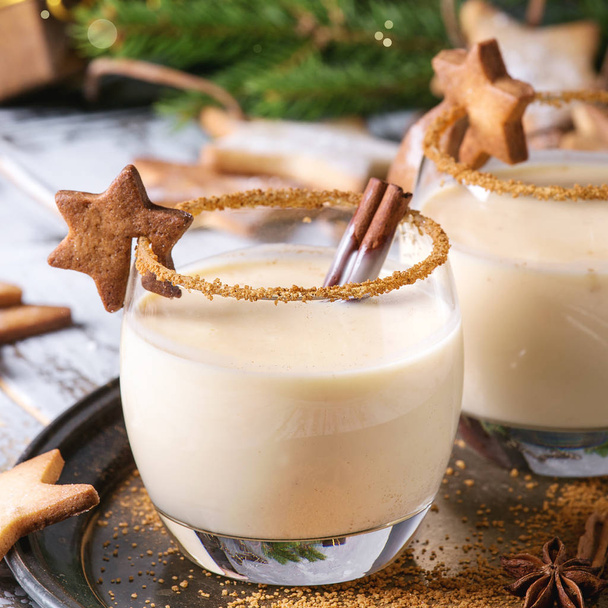 Eggnog Christmas milk cocktail with cinnamon, served in two glasses on vintage tray with shortbread star shape sugar cookies, decor toys, fir branch over white wooden plank table. Close up. Square image - Fotó, kép
