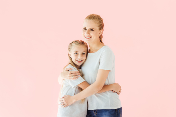 beautiful mother and daughter in white t-shirts embracing and looking at camera isolated on pink - Foto, Imagem