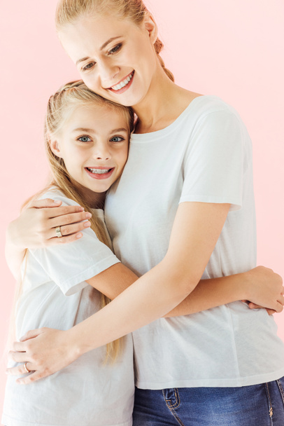 ευτυχισμένη μητέρα και κόρη σε λευκά μπλουζάκια Αγκαλιάζοντας και βλέπουν τα φωτογραφικών μηχανών που απομονώνονται σε ροζ - Φωτογραφία, εικόνα