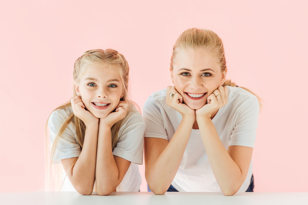 счастливые молодые мать и дочь в белых футболках опираясь на столешницу и глядя на камеру изолированы на розовый
 - Фото, изображение