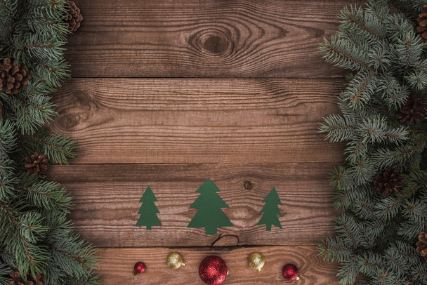 top view of green paper fir trees with shiny baubles and coniferous branches with pine cones on wooden surface, christmas background - Foto, Imagen