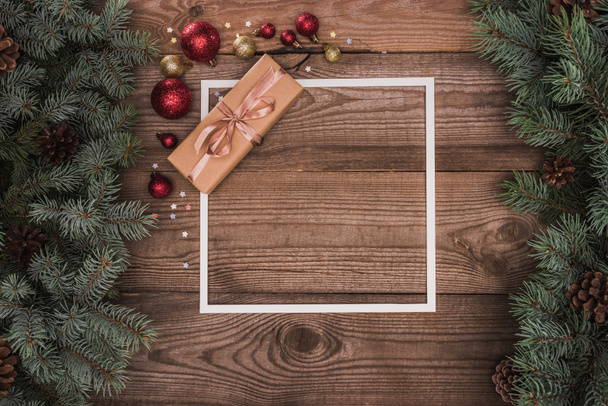 bovenaanzicht van witte frame, kerstcadeau en glanzende kerstballen op houten oppervlak met spar twijgen en dennenappels - Foto, afbeelding