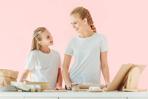 smiling mother and daughter in white t-shirts looking at each other while cooking together isolated on pink - Photo, Image