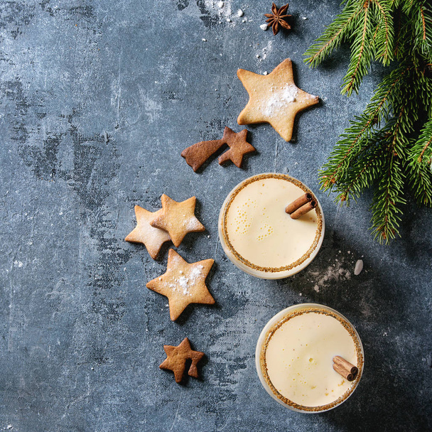 Eggnog Christmas milk cocktail with cinnamon, served in two glasses with shortbread star shape sugar cookies different size, fir branch over blue texture background. Top view with space. Square image - Photo, Image