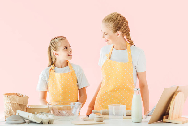 glückliche Mutter und Tochter schauen einander beim Zubereiten von Teig isoliert auf rosa - Foto, Bild
