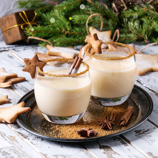 Eggnog Christmas milk cocktail with cinnamon, served in two glasses on vintage tray with shortbread star shape sugar cookies, decor toys, fir branch over white wooden plank table. Square image - Foto, afbeelding