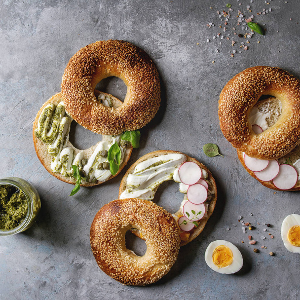 Variety of homemade bagels with sesame seeds, cream cheese, pesto sauce, eggs, radish, herbs with ingredients above over grey texture background. Top view, space. Square image - Photo, Image