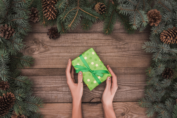 partial top view of person holding christmas present above wooden surface with coniferous branches and pine cones - Фото, зображення