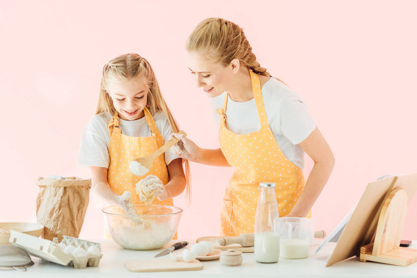 happy young mother and adorable little daughter in yellow aprons making dough together isolated on pink - Foto, afbeelding
